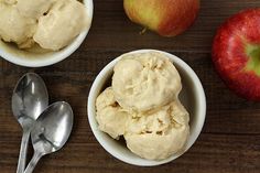 two bowls filled with ice cream next to an apple and spoons on a wooden table