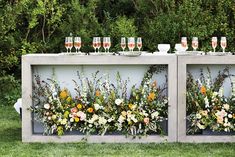 an outdoor bar with flowers and wine glasses on the top, surrounded by greenery