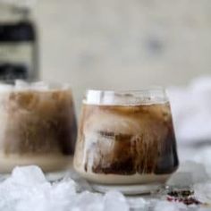 two glasses filled with iced coffee sitting on top of snow covered ground next to a bottle