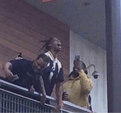 two men are standing on the side of a building and one is holding up his cell phone