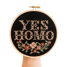 a hand holding up a cross stitch hoop with the words yes, he is home on it