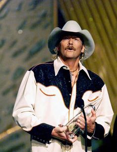 a man wearing a cowboy hat and holding a glass in his hand while standing next to a microphone