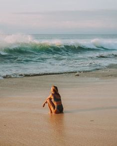 a woman sitting on top of a sandy beach next to the ocean with waves coming in