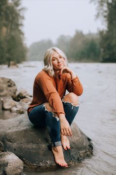 a woman sitting on top of a rock next to a river with trees in the background