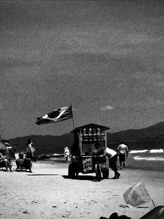 an old truck parked on the beach with people walking around it and flags flying in the wind