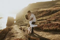 a man and woman are hugging on the rocks by the water at sunset or sunrise