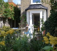 a house that is surrounded by plants and flowers in front of the door, with an open window