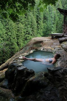 a woman laying in a pool of water next to some rocks and trees on the side of a cliff