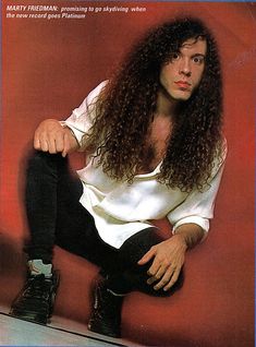 a man with long curly hair sitting on top of a skateboard in front of a red wall