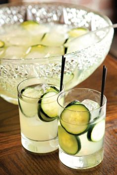 two glasses filled with cucumbers sitting on top of a wooden table next to a bowl