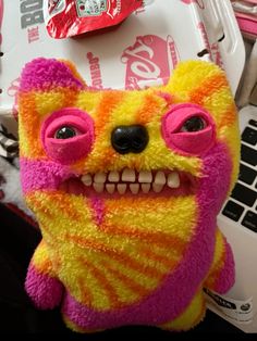 a teddy bear with pink and yellow eyes sitting in front of a laptop