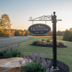 the sign for true stone is in front of some purple flowers and grass with trees behind it