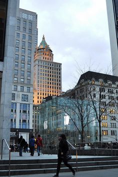 people are walking on the steps in front of tall buildings at dusk, with an apple store lit up behind them