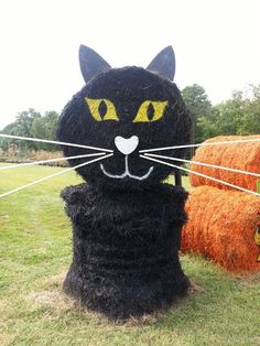 a black cat made out of hay sitting on top of a grass field next to bales of hay