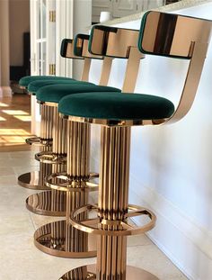 three gold barstools with green velvet seats in front of a white wall and marble floor