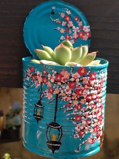 a blue tin can with succulents and flowers painted on it, hanging from a wooden fence