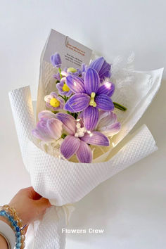 a hand holding a bouquet of purple and yellow flowers with white paper wrapping around it