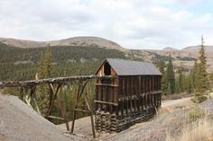 an old wooden building sitting on top of a hill in the middle of trees and mountains