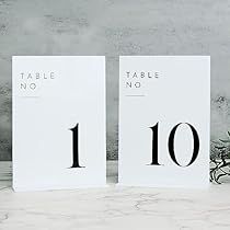 two white tables with numbers on them in front of a vase filled with flowers and greenery