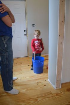 a young boy standing next to a blue bucket on top of a hard wood floor