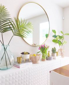 a white table topped with a mirror and vases filled with green plants next to a wooden table