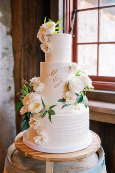 a wedding cake with white flowers on top sits on a barrel in front of a window