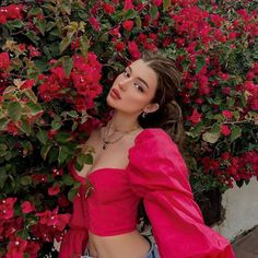 a woman in pink top and jeans standing next to red flowered bush with her hands on her hips