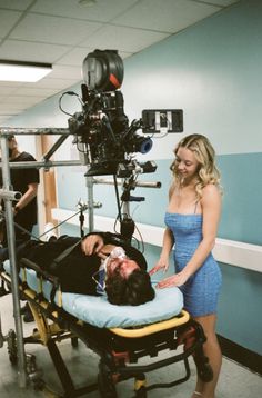 a woman standing next to a man laying on top of a bed in front of a camera