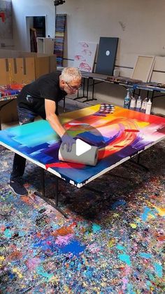 an older man is painting on a table in the middle of a room filled with paintings