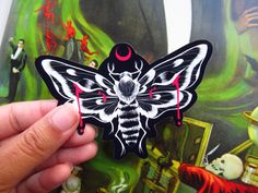 a hand holding up a sticker with a moth on it's face in front of a painting