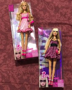 two barbie dolls in their packaging on a purple and pink patterned clothed bed spread