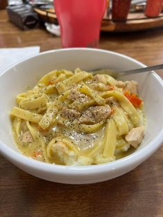 a white bowl filled with pasta and meat on top of a wooden table next to a red cup