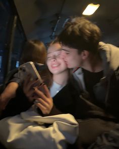 two people sitting on a bus looking at a cell phone and kissing each other's foreheads