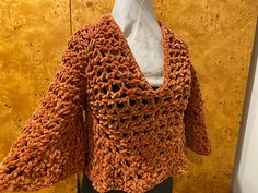 an orange crocheted jacket on display in front of a yellow wall with a white shirt underneath it