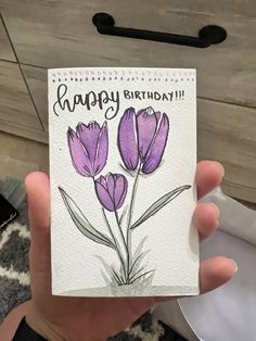 a hand holding up a card with purple flowers on it and the words happy birthday written in cursive writing