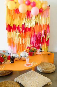 an orange table topped with lots of balloons next to a wall covered in streamers