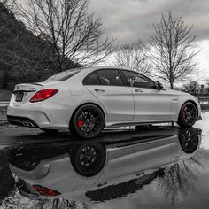 a white car parked in front of a mirror on the ground with trees behind it