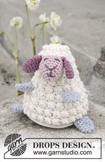 a crocheted sheep sitting on top of a cement slab with flowers in the background