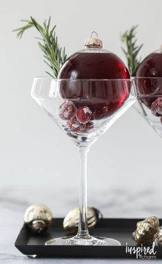 two wine glasses filled with red liquid and christmas ornaments on a black tray next to silver balls