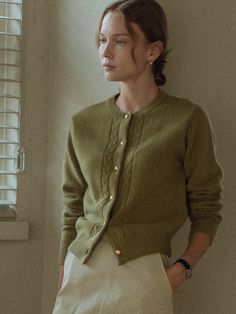 a woman standing in front of a window wearing a green sweater and white skirt with buttons