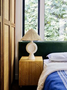 a bed sitting under a window next to a lamp on top of a wooden table