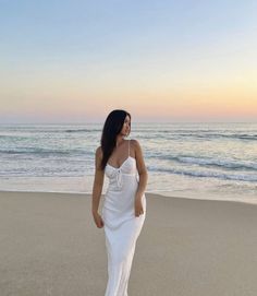 a woman in a white dress standing on the beach at sunset with her back to the camera