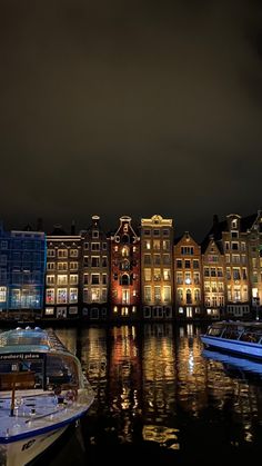 several boats are docked in the water near some buildings at night time, with lights reflecting on the water