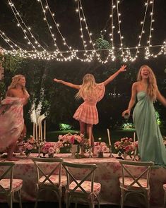 three women in dresses standing on a table with lights strung above them and flowers all around