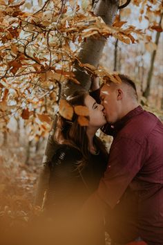 a man and woman are kissing under a tree in the fall leaves with their faces close to each other