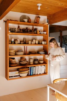 a woman standing in front of a wooden shelf filled with bowls and plates on it