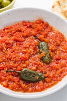 a white bowl filled with tomato sauce next to some pita bread and green peppers