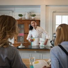 a woman standing in front of a counter next to another woman holding a book and smiling