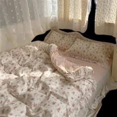 an unmade bed in front of two windows with white curtains and pink flowers on it