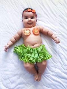 a baby wearing a green skirt and orange slices on it's chest, laying on a white sheet